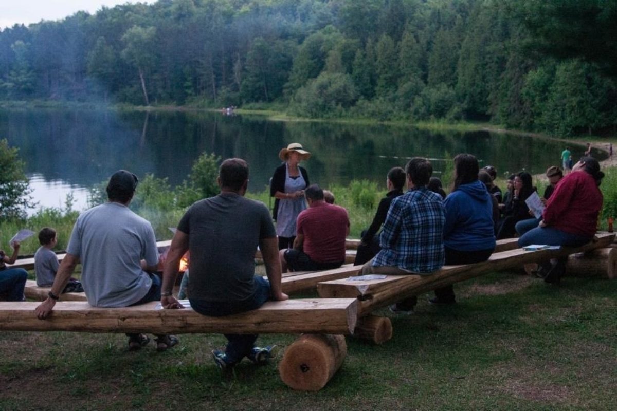 Des gens rassemblés sur des bancs autour d’un feu de camp près de la rive d’un lac.